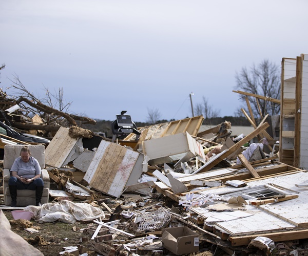 a woman sits in a big recliner chair amongst the ruble of her home.