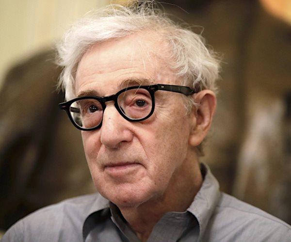 woody allen stands and listens wearing horn-rimmed glasses